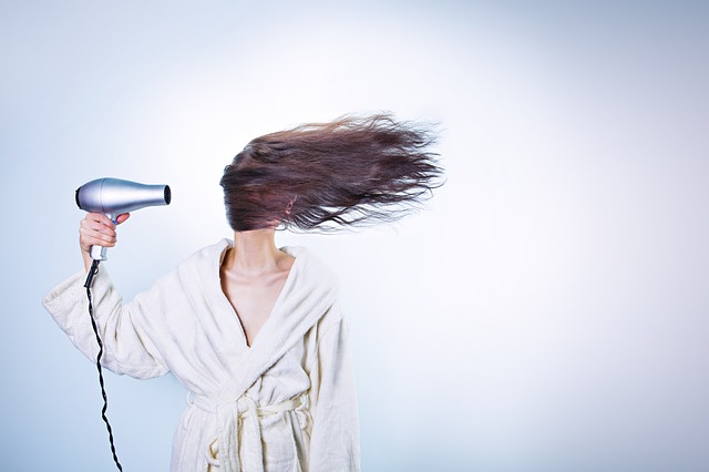 Long haired brunette woman stood to the left hand side of a white background. She is wearing a cream dresing gown and holding a silver hairdryer on her right hand. the hair dryer is blowing her hair all across her face so you can't see her face at all